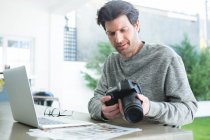 Homme utilisant un ordinateur portable et tenant un appareil photo numérique à la maison — Photo de stock