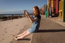 Sorrindo jovem mulher tirando foto do mar com telefone celular na praia — Fotografia de Stock
