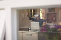 Старший чоловік використовує гарнітуру віртуальної реальності з ноутбуком на кухні вдома — стокове фото
