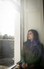 Nachdenkliche Frau, die durch Fenster schaut, während sie zu Hause auf Fensterbank sitzt — Stockfoto