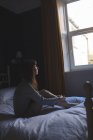 Femme regardant par la fenêtre tout en étant assis sur le lit à la maison — Photo de stock