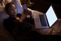 Mujer usando el teléfono móvil mientras se relaja en la cama en el hotel - foto de stock