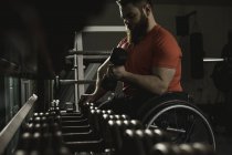 Handicapped man on wheelchair lifting dumbbell from rack in gym — Stock Photo