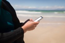 Середина дівчини в гідрокостюмі з використанням мобільного телефону на пляжі — стокове фото