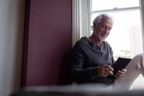 Старший чоловік використовує цифровий планшет біля вікна у вітальні вдома — стокове фото