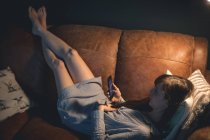 Жінка використовує мобільний телефон, лежачи на дивані у вітальні — стокове фото