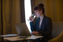 Empresária usando laptop enquanto bebe vinho no quarto de hotel — Fotografia de Stock
