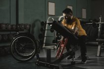 Man sitting on bench press in gym by wheelchair — Stock Photo