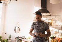 Homem segurando xícara de café e smartphone na cozinha em casa . — Fotografia de Stock