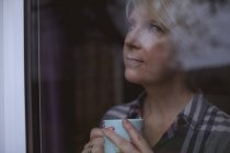 Femme mûre réfléchie tenant tasse de café tout en regardant par la fenêtre à la maison — Photo de stock