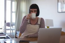 Junge Frau trinkt Kaffee, während sie zu Hause Laptop benutzt — Stockfoto
