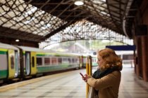Mujer joven pelirroja apoyada en la barandilla usando su teléfono móvil en la plataforma ferroviaria - foto de stock