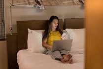 Donna d'affari seduta sul letto con il telefono mentre lavora sul computer portatile in hotel — Foto stock