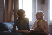 Дочь дарит старшей женщине цветы, сидя на диване в гостиной дома — стоковое фото