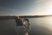 Mulher tirando foto com celular perto da ribeira durante o pôr do sol . — Fotografia de Stock