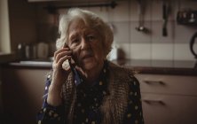 Mujer mayor preocupada hablando por teléfono en la cocina en casa - foto de stock