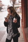 Menina bonita usando celular enquanto toma café fora do shopping — Fotografia de Stock