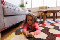 Девушка лежит на ковре и с помощью мобильного телефона в гостиной дома — стоковое фото