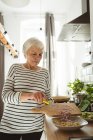 Старша жінка додає нарізану цибулю в миску на кухні — стокове фото