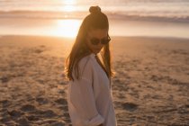 Frau mit Sonnenbrille steht bei Sonnenuntergang am Strand. — Stockfoto