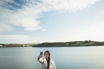 Frau macht bei Sonnenuntergang Selfie mit Handy in Flussnähe. — Stockfoto
