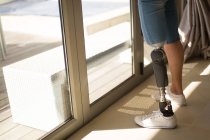 Sección baja de la mujer con la pierna protésica de pie cerca de la ventana en casa . - foto de stock