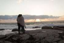 Rückansicht eines in Schaufel gehüllten Paares, das während des Sonnenuntergangs auf einem Felsen nahe der Küste steht — Stockfoto