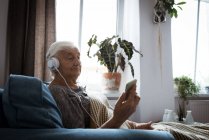 Старша жінка слухає музику на навушниках, використовуючи мобільний телефон у вітальні — стокове фото