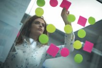 Female executive sticking adhesive notes on glass wall in office — Stock Photo
