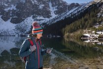 Туристка с помощью мобильного телефона на берегу озера в зимний период — стоковое фото