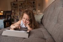 Улыбающаяся девочка лежит на диване и пользуется цифровым планшетом в гостиной дома — стоковое фото