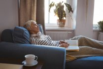 Seniorin schläft auf dem Sofa, während sie zu Hause im Wohnzimmer das Buch liest — Stockfoto