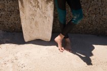 Petite fille en combinaison debout avec planche de surf sur la plage — Photo de stock