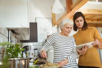 Дочь показывает пожилой женщине рецепт на таблетке на кухне во время приготовления пищи — стоковое фото