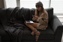 Mulher de chapéu de lã usando telefone celular no sofá na sala de estar em casa . — Fotografia de Stock