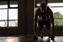 Young man with long red hair doing push-ups with dumbbells in fitness studio. — Stock Photo