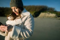 Junge Frau mit Wollmütze benutzt Handy am Flussufer. — Stockfoto