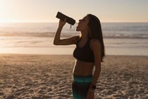 Atleta feminina bebendo água na praia ao entardecer . — Fotografia de Stock