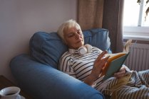 Старша жінка розслабляється на дивані, читаючи книгу у вітальні вдома — стокове фото