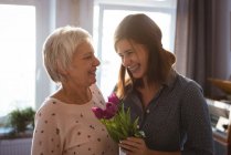Daughter giving senior woman smiling while holding flowers in living room at home — Stock Photo
