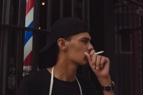 Jeune coiffeur fumant de la cigarette à l'entrée de son magasin — Photo de stock