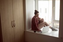 Mulher com café ao usar laptop no peitoril da janela em casa . — Fotografia de Stock