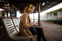 Mujer joven listando música mientras usa su teléfono móvil en la plataforma ferroviaria - foto de stock