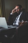 Homme d'affaires réfléchi utilisant un ordinateur portable tout en ayant whisky à la chambre d'hôtel — Photo de stock