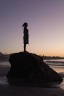 Silueta de mujer de pie sobre roca en la playa al atardecer . - foto de stock