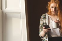 Frau trinkt Kaffee, während sie ihr Handy zu Hause benutzt — Stockfoto