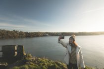 Femme prenant selfie avec téléphone portable près de la rivière pendant le coucher du soleil . — Photo de stock