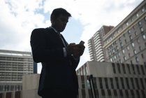 Vista lateral del hombre de negocios utilizando el teléfono móvil contra rascacielos - foto de stock