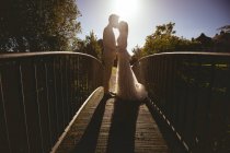 Жених и невеста целуются на пешеходном мосту в саду в солнечный день — стоковое фото