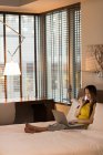 Donna d'affari seduta sul letto con il telefono mentre lavora sul computer portatile in hotel — Foto stock